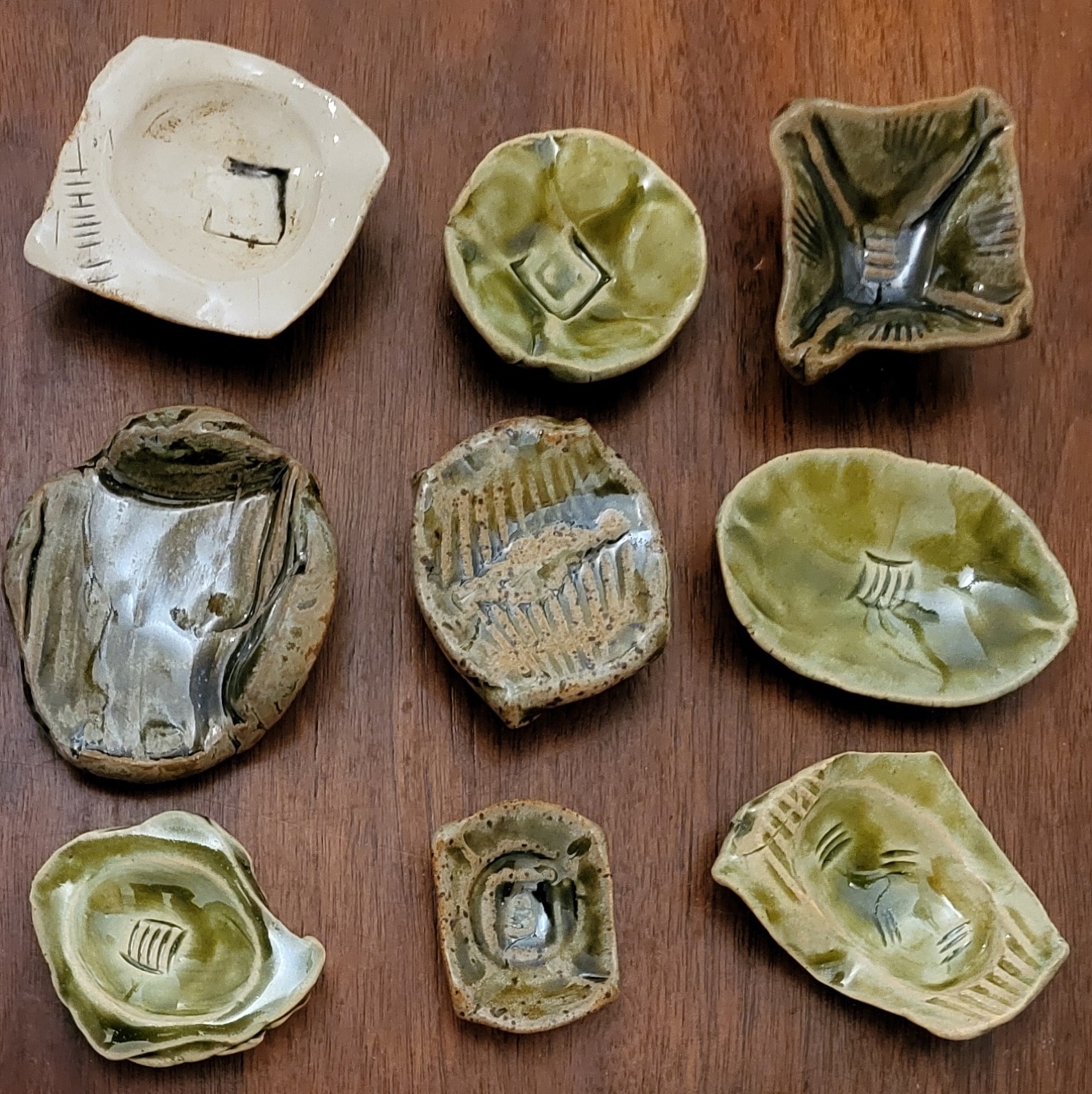 A grid of nine miniature ceramic forms, each about an inch wide, in greens, ivories and earth tones with carved interior details.