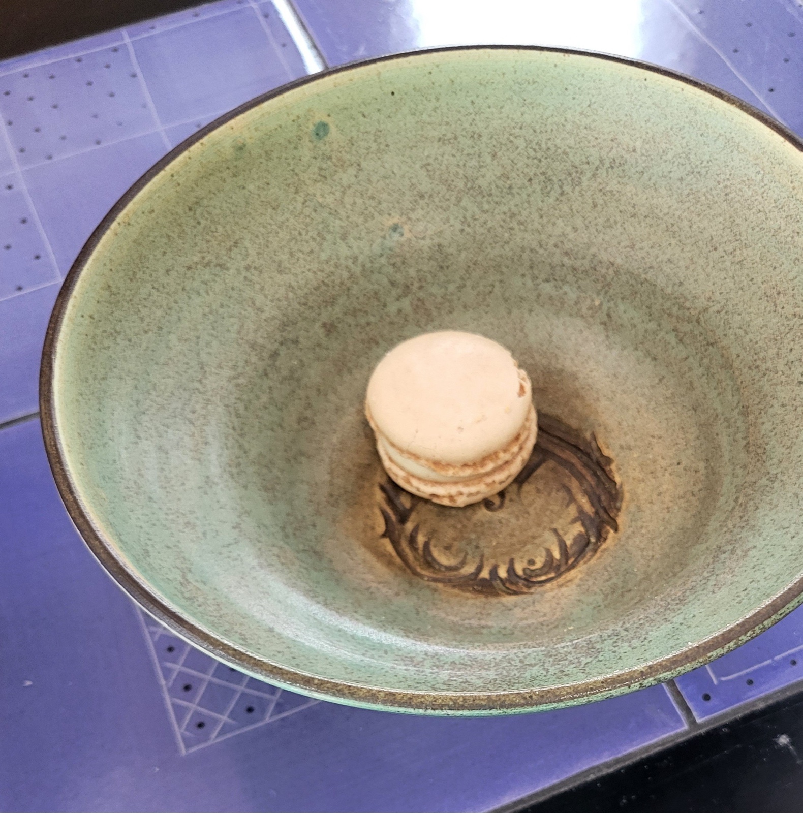 A teal-green stoneware bowl with a scrolled medallion impression at its center, holding a single macaron.