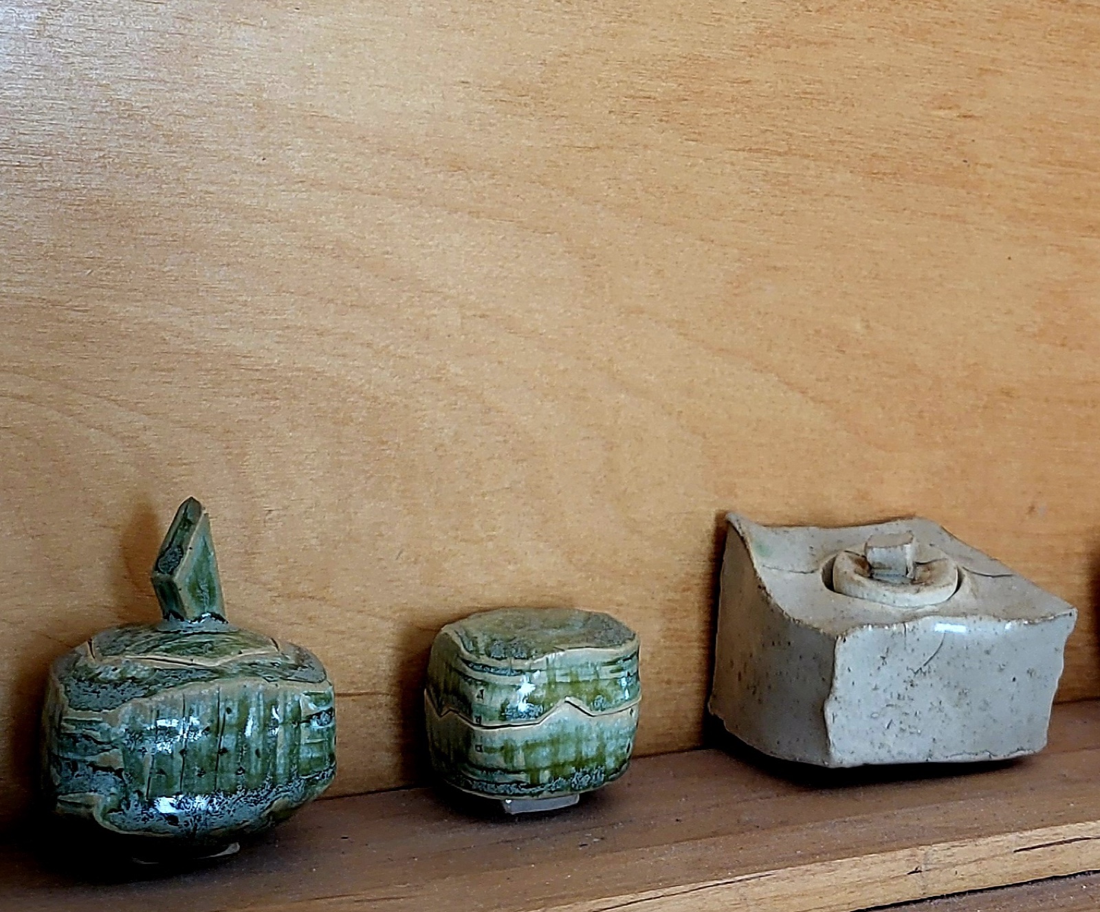 Three small lidded ceramic vessels on a wooden shelf: two with green glaze, one square in cream.