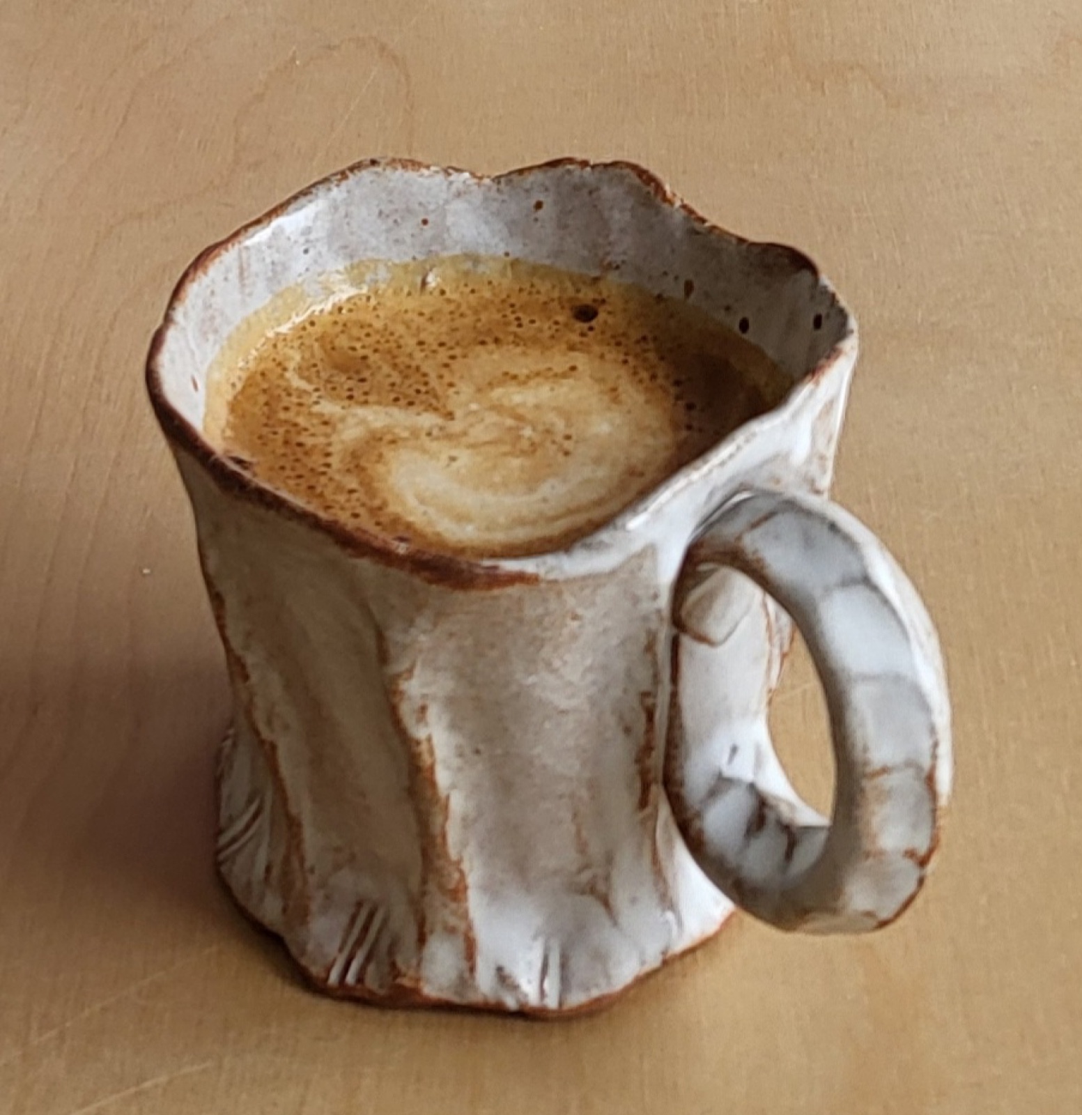 A pinched stoneware coffee mug in a white glaze warming to earth tones at the edges, holding espresso, on a wood surface.